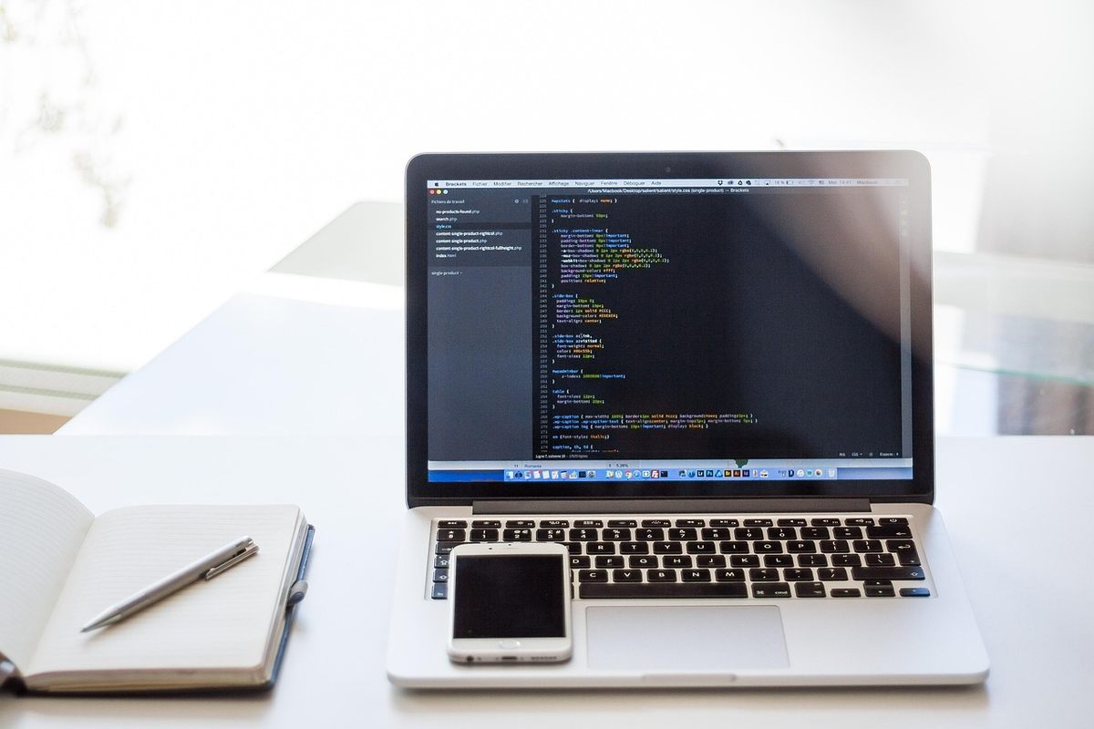 Laptop displaying code, smartphone on keyboard, notebook with pen, bright window