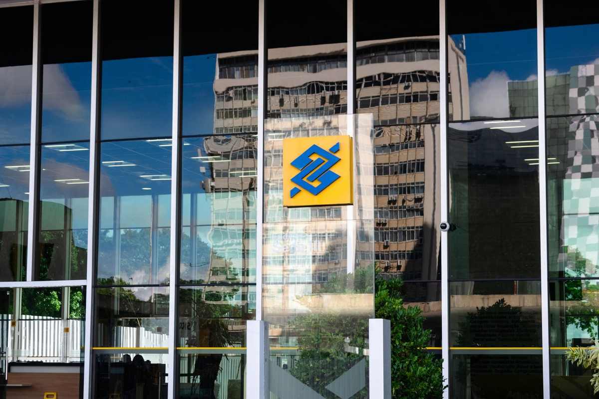 Façade vitrée d'un bâtiment avec logo bleu sur fond jaune, reflet d'un gratte-ciel et du ciel
