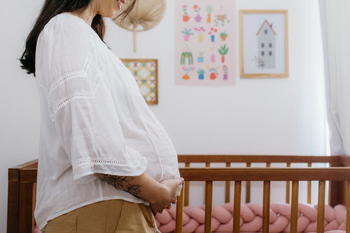 Femme enceinte dans une chambre, main sur ventre, lit d'enfant avec coussin tressé, oeuvres murales